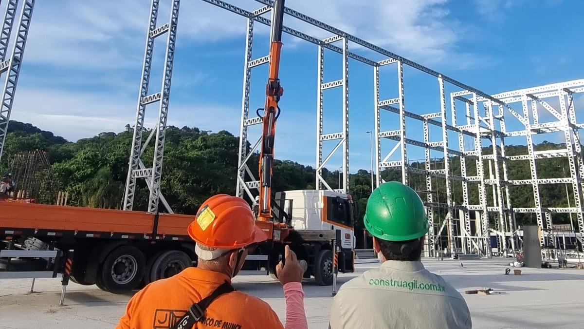 Dois técnicos com capacete acompanham a montagem de uma estrutura em light steel
              frame, com a grua a posicionar painéis ao fundo.